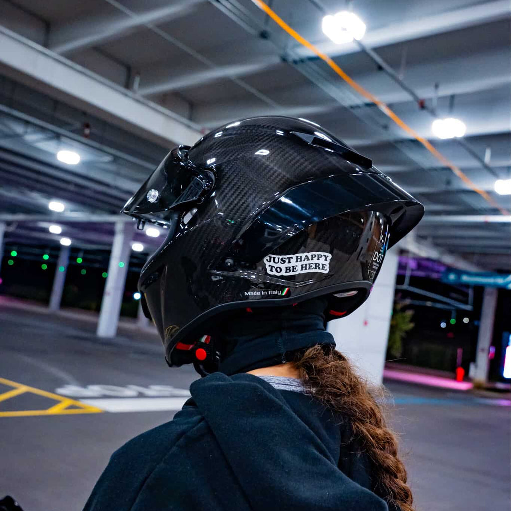 A person wearing a glossy black carbon-fiber full-face motorcycle helmet with a clear visor and a white sticker that reads 'JUST HAPPY TO BE HERE', photographed from behind in an indoor parking structure.