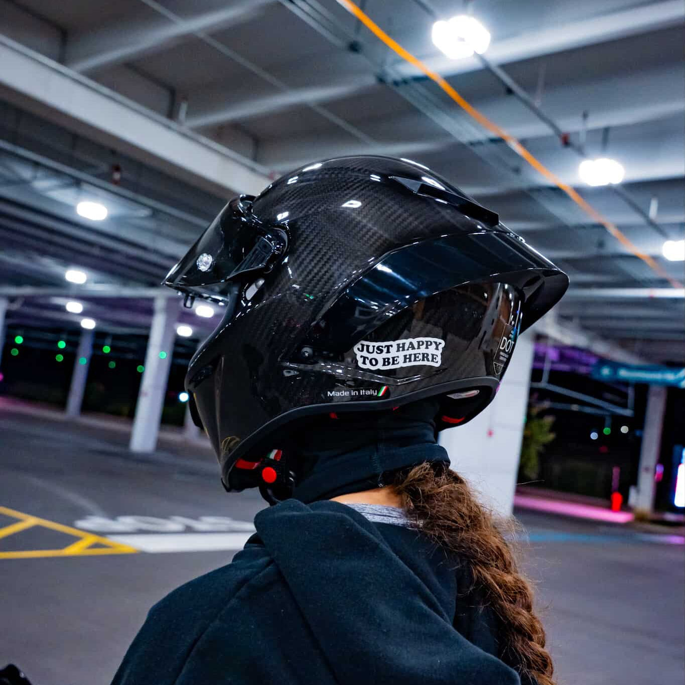 A person wearing a glossy black carbon-fiber full-face motorcycle helmet with a clear visor and a white sticker that reads 'JUST HAPPY TO BE HERE', photographed from behind in an indoor parking structure.